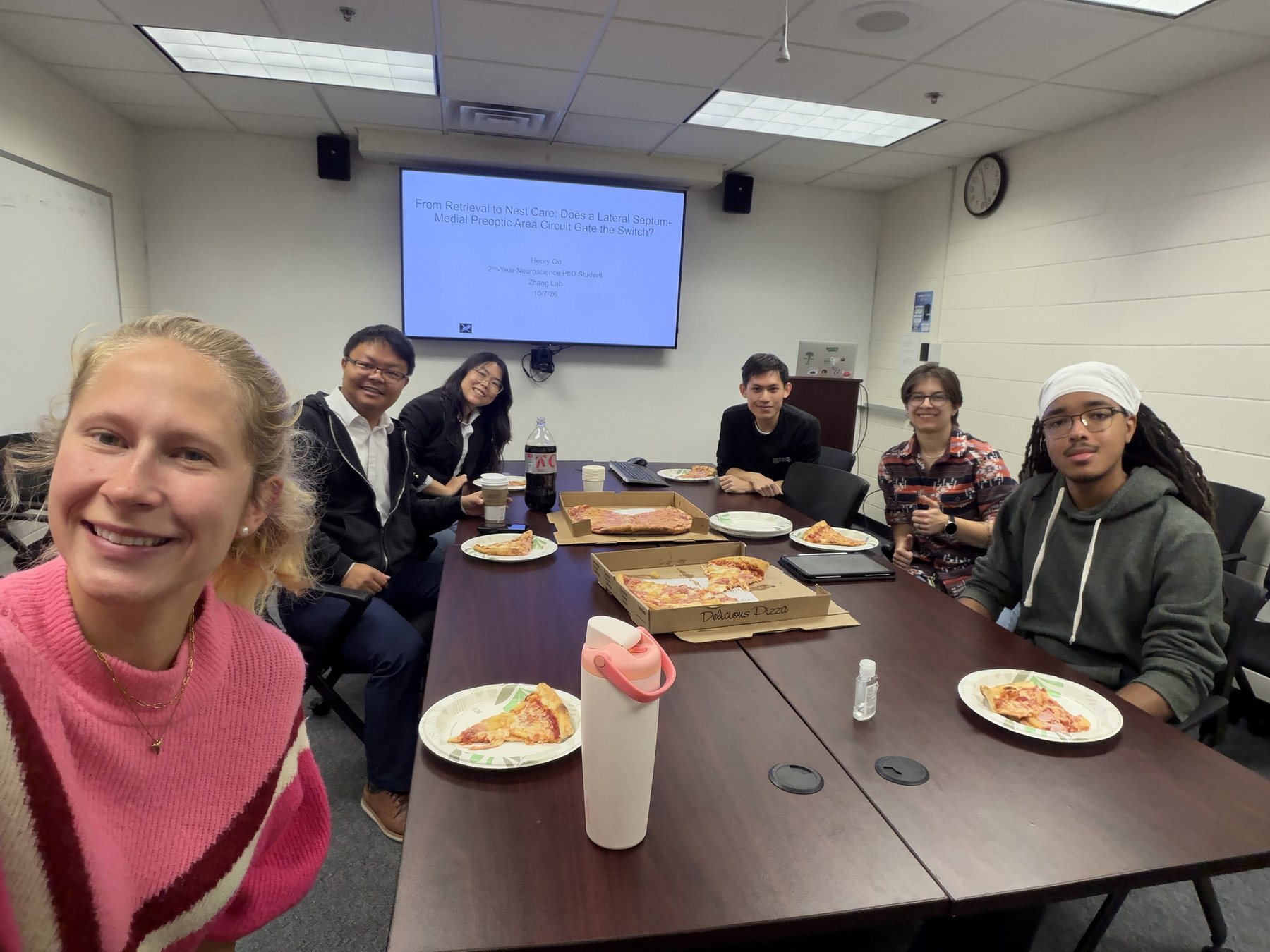 The Zhang Lab around a table with pizza at group study, with Henry Oo's title slide on the screen, October 7, 2026